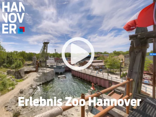 Szenerie im Erlebnis-Zoo Hannover mit Wasser, Felsen, Schiff und Westernstadt-Kulisse unter blauem Himmel.